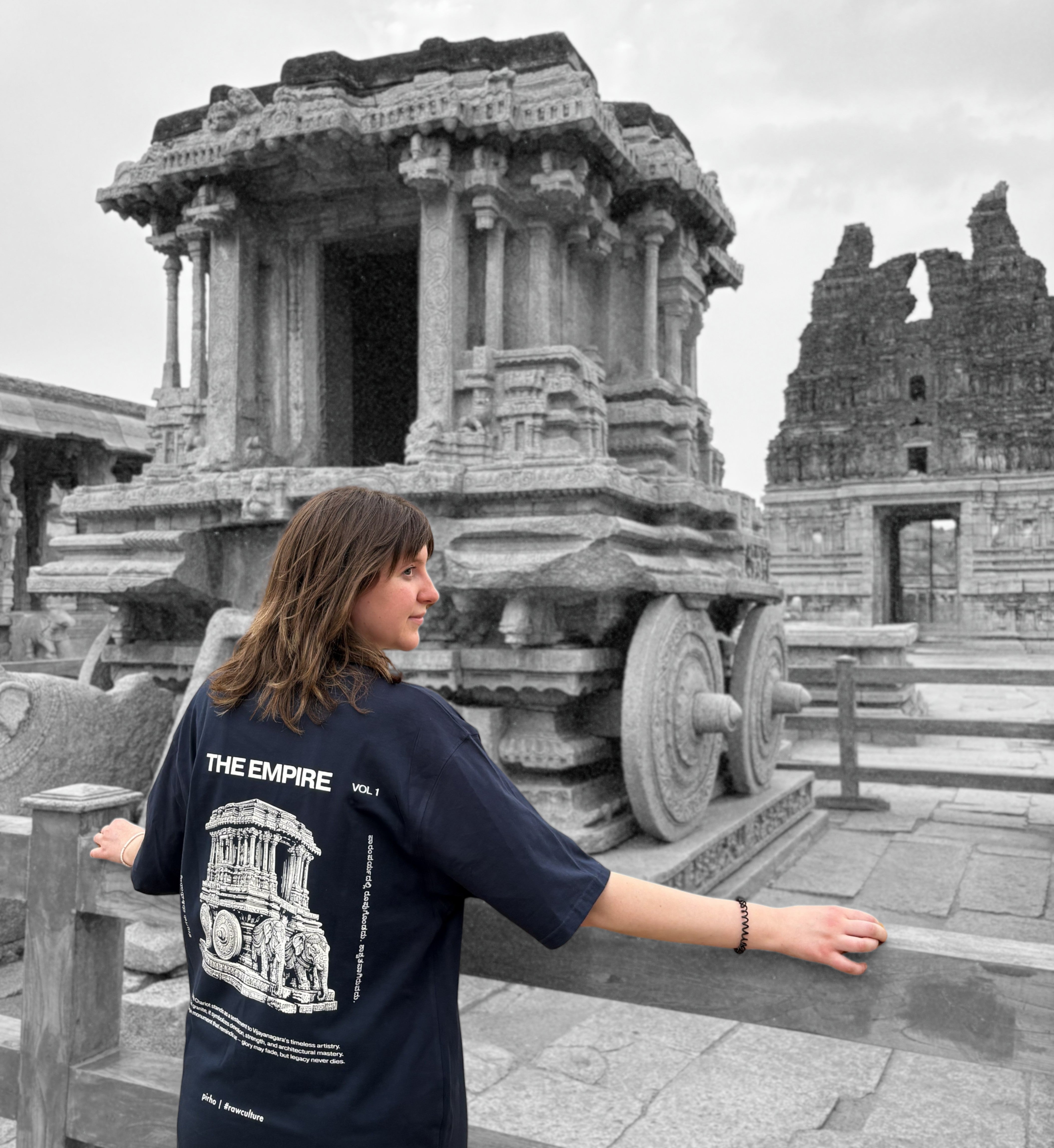 Person wearing a black t-shirt with a design and text, standing in front of an ancient stone chariot.